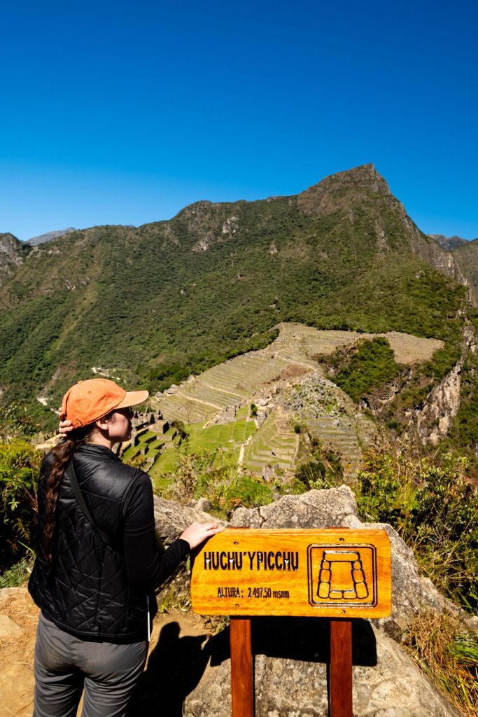 A visitor beside the Huchu'ypicchu sign at Machu Picchu, with Inca terraces and Huayna Picchu rising behind